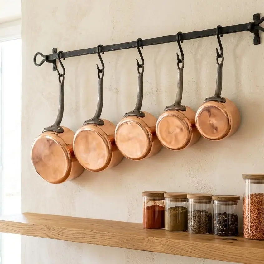 Five-piece Moroccan copper cookware set hanging from a black wrought iron wall-mounted rack, showing the shiny round copper pot bases above organized spice jars.