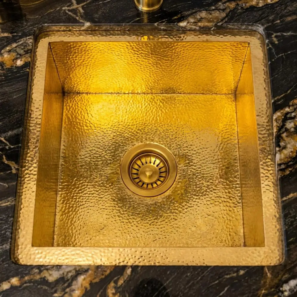 Top-down view of a square hammered brass kitchen sink showcasing intricate textured patterns and a center drain on a black marble countertop.
