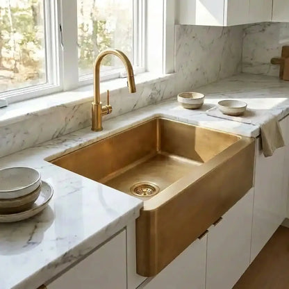 Close up view of a 30 inch unlacquered brass farmhouse kitchen sink embedded in white veined marble countertops next to a bright window.