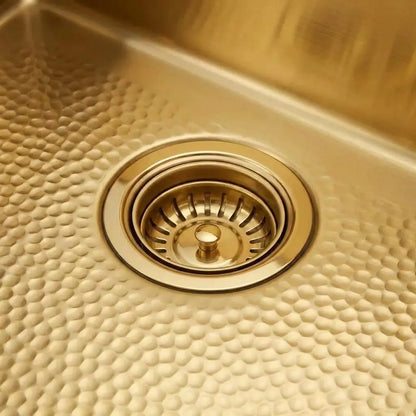 Close-up of a 30-inch unlaquered brass kitchen sink showing the standard 3.5-inch drain opening with matching basket strainer and scratch-resistant hand-hammered texture