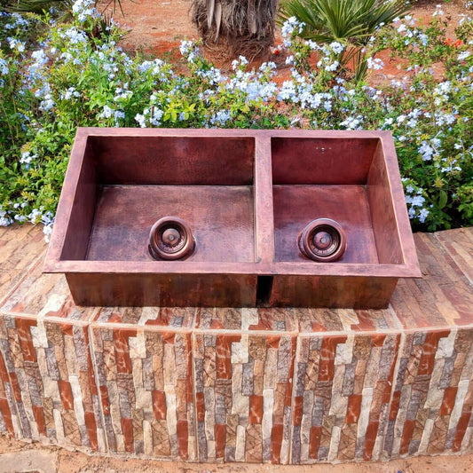 Handmade hammered double copper sink with two compartments and drains, set on a stone surface amidst garden flowers.