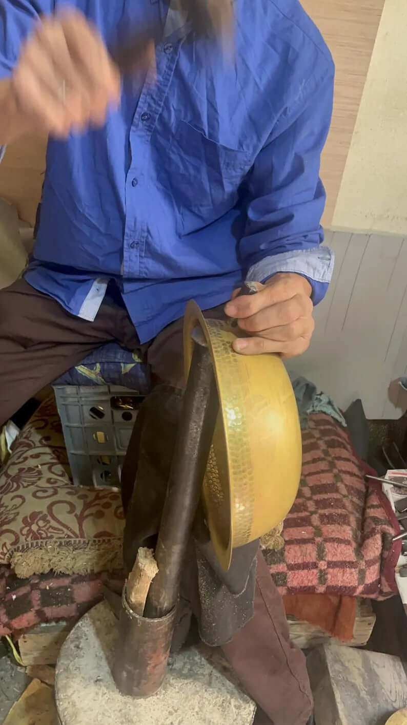 Artisan shaping a brass sink with a hammer in a workshop, showcasing traditional craftsmanship.
