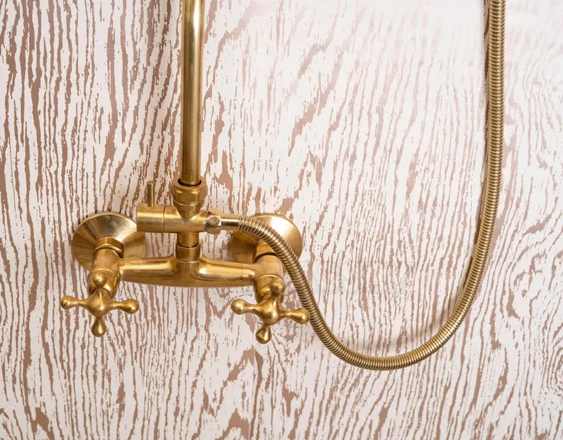 Unlacquered brass shower system with dual handles and handheld sprayer against a textured wall.
