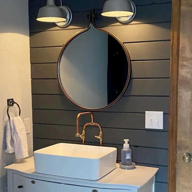 Handmade copper faucet above white vessel sink in stylish bathroom with dark wood paneling and round mirror.