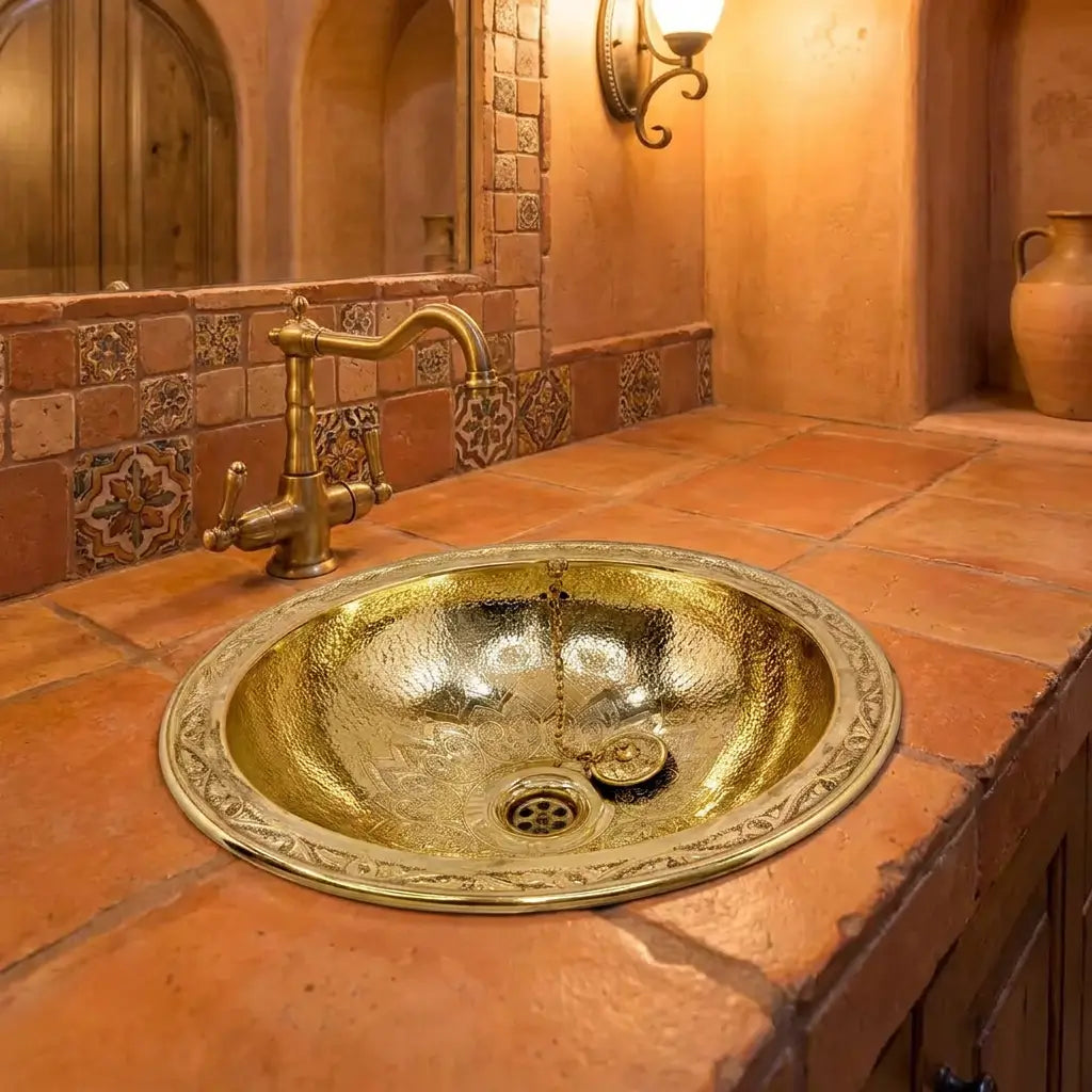 Round hand-hammered brass drop-in sink with engraved wide rim paired with an antique brass bridge faucet on a rustic terracotta tile vanity
