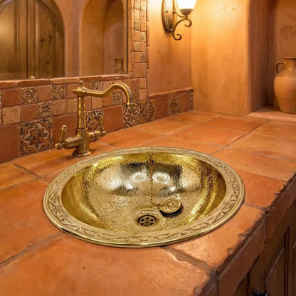 Round hand-hammered brass drop-in sink with engraved wide rim paired with an antique brass bridge faucet on a rustic terracotta tile vanity