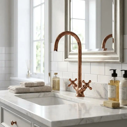 Antique copper gooseneck faucet on a white marble vanity sink with a silver mirror frame and tile wall background.