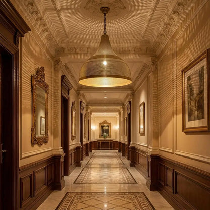 An illuminated antique brass dome chandelier hanging in a classic residential hallway, casting complex grid-like light patterns on Victorian-style wood wall paneling and framed artwork.
