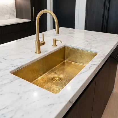Close-up view of a golden brass undermount sink on a white marble kitchen island with an elegant arched gold faucet