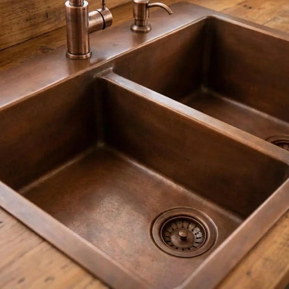 Handmade double bowl copper kitchen sink with a textured hammered finish, showcasing an antique farmhouse basin design installed on a wooden countertop next to a matching brass faucet and drain strainer.