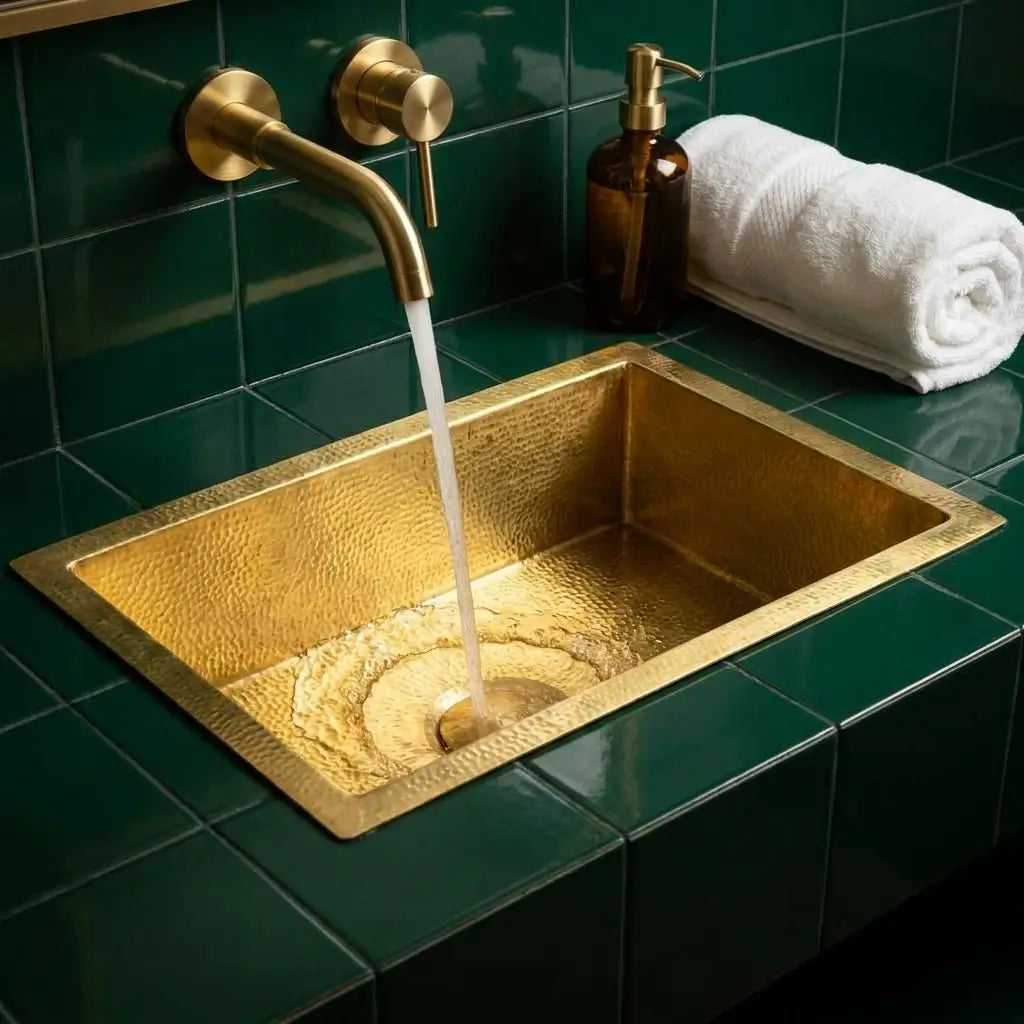 Golden brass bathroom sink integrated into emerald green tiles with water flowing from a wall-mounted gold faucet next to a rolled white towel