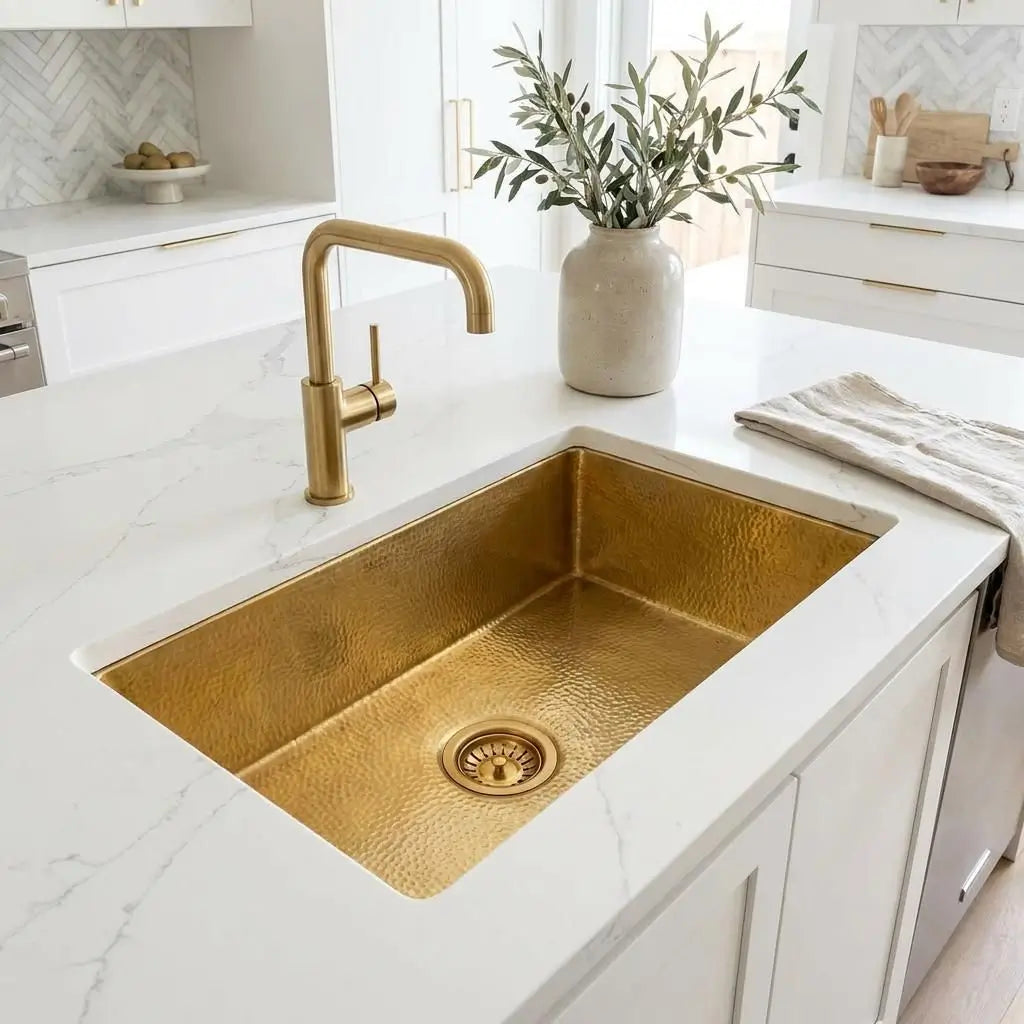 Angled high view of a golden brass undermount sink built into a bright white countertop featuring a gold faucet and a rustic olive branch vase