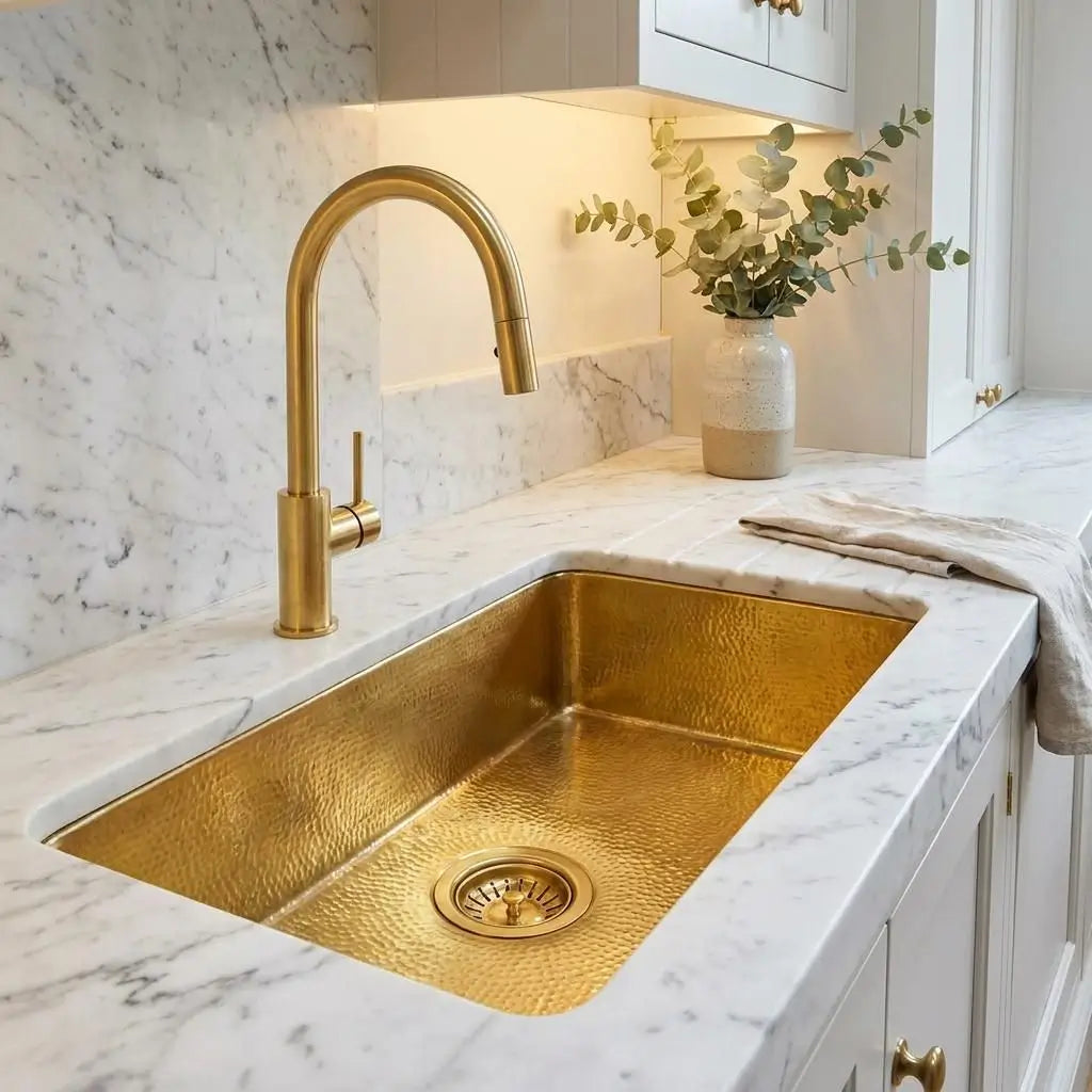 Golden brass undermount bathroom sink integrated into a white marble countertop with grey veining featuring a tall gold faucet and a decorative vase