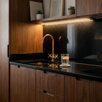 Handmade brass Moroccan vanity tap with gooseneck spout installed on a dark countertop in a modern wooden kitchen with warm under-cabinet lighting.