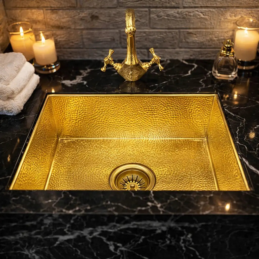 Square hammered brass Moroccan sink undermounted in a black marble countertop, styled with a gold traditional faucet and lit candles.