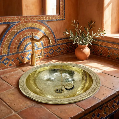 Hand-hammered polished brass drop-in washbasin with engraved rim and gooseneck faucet embedded in a rustic terracotta tiled countertop with Moroccan mosaic zellige backsplash