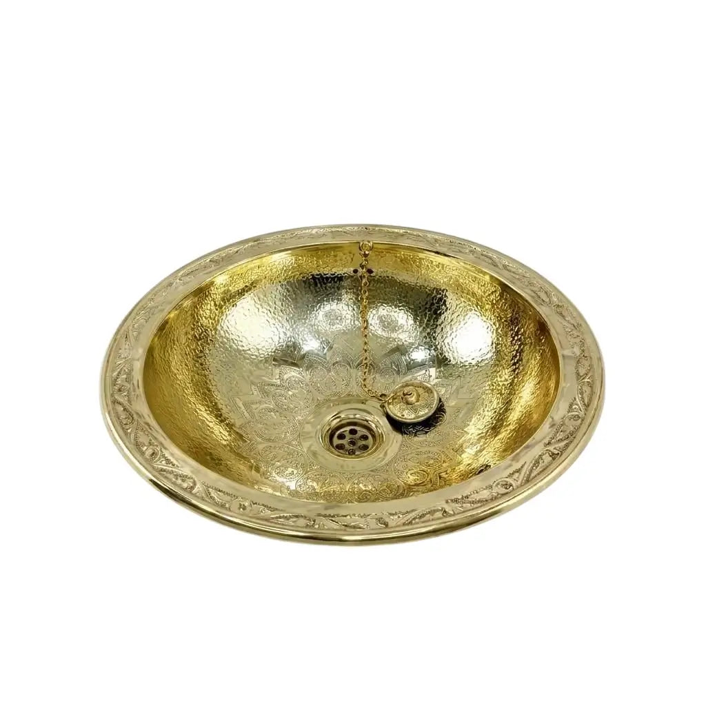 Oval hand-hammered brass drop-in basin featuring intricate floral engraving on the interior bowl and a wide patterned rim with integrated drain plug