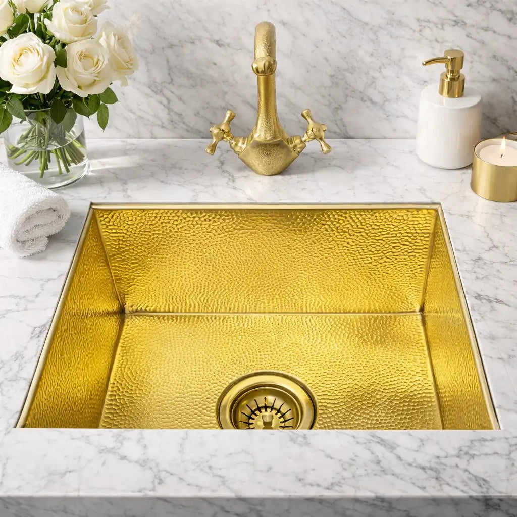 Top view of a custom handcrafted brass Moroccan sink installed under a marble countertop, featuring a gold brass faucet, drain strainer, and decorative bathroom accessories.