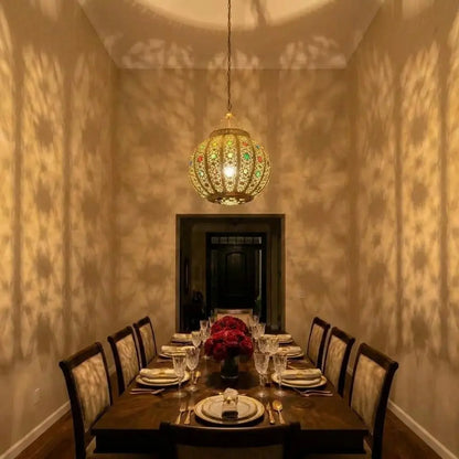 Handmade brass lampshade with colorful glass accents hanging in a long narrow dining room, casting precise geometric patterns of light and shadows on the side walls.