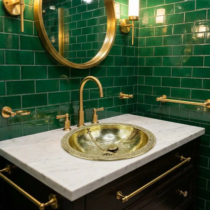 Hand-hammered round brass vessel sink installed on a white countertop against a green tiled bathroom wall with a matching gold gooseneck faucet.