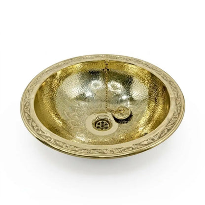 Studio shot of an oval hand-hammered brass vessel sink with a floral engraved flat rim and an included drainage plug on a plain white background.