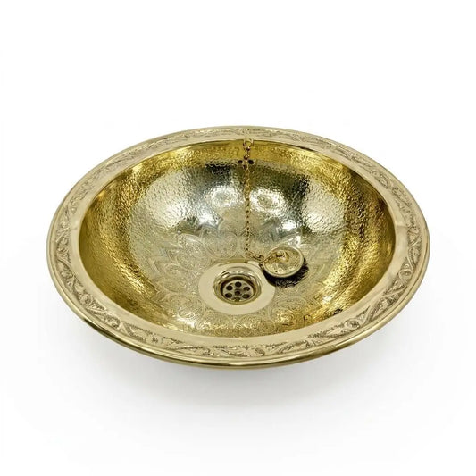 Studio shot of an oval hand-hammered brass vessel sink with a floral engraved flat rim and an included drainage plug on a plain white background.