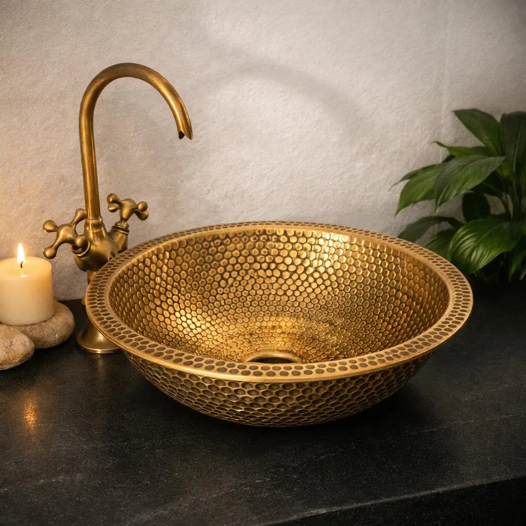 Modern custom made brushed brass bathroom sink with a detailed honeycomb hammered finish, styled with a left-side antique faucet, lighting candle, and green plant on a premium dark charcoal countertop.