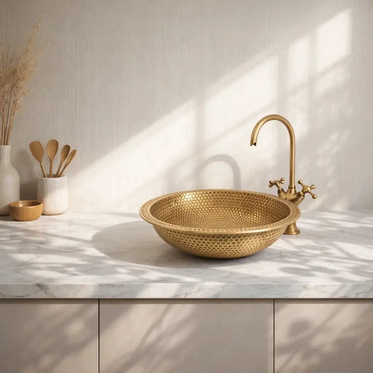 Modern custom made brushed brass bathroom sink with a hammered finish, styled with an antique brass faucet on a luxurious white marble countertop with natural sunlight.