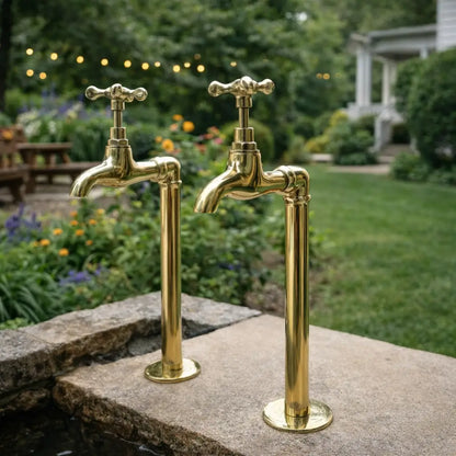 A pair of unlacquered brass water faucets with cross handles installed on a stone ledge in a backyard garden setting with string lights and green lawn in the background.