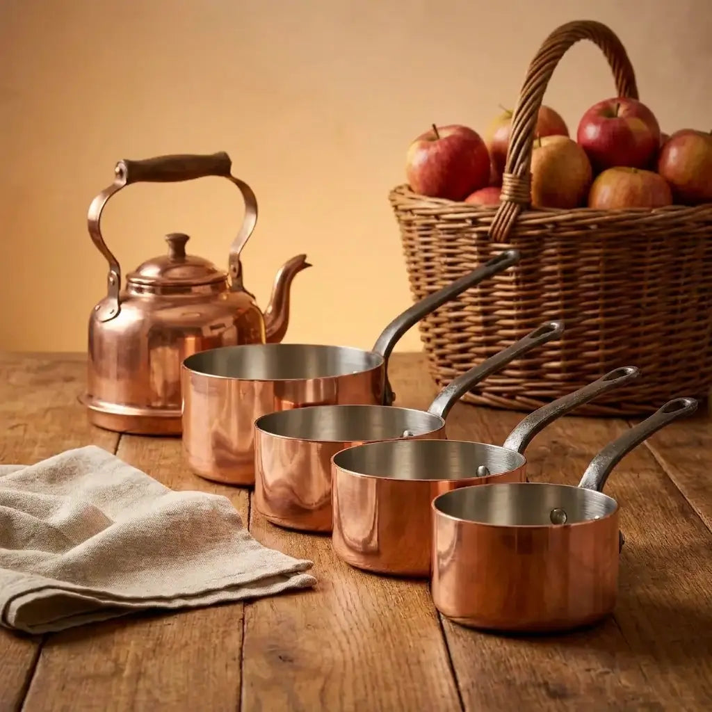 Four graduated Moroccan copper saucepans alongside a vintage copper tea kettle on a rustic wooden table, featuring an apple basket and traditional tin-lined interiors.