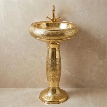 Artisanal Moroccan engraved brass pedestal washbasin with a matching high-arc brass faucet, displaying detailed ethnic patterns against a neutral textured studio backdrop.