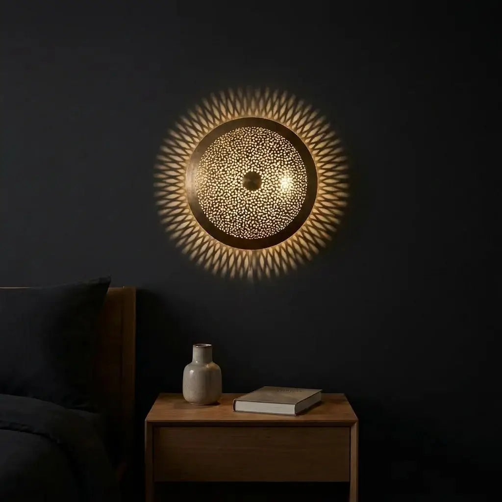 Moroccan wall light displaying handmade artisan brass lighting with a warm glowing sunburst reflection on a dark wall above a wooden nightstand.