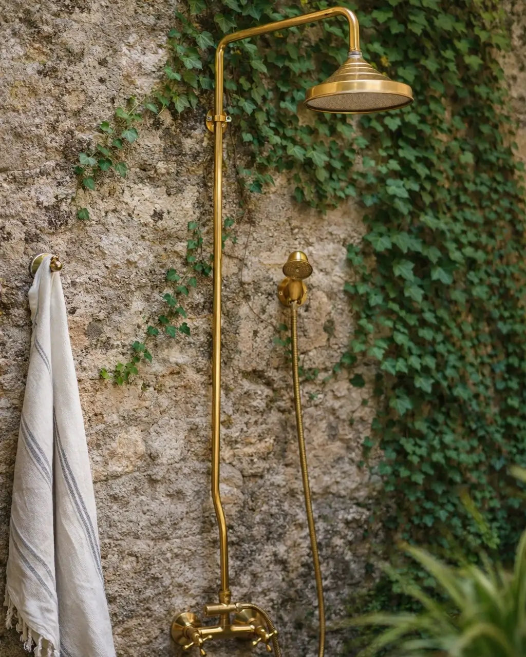 Outdoor unlacquered brass shower system with a Moroccan rain head mounted on a rustic stone wall with green ivy vines.