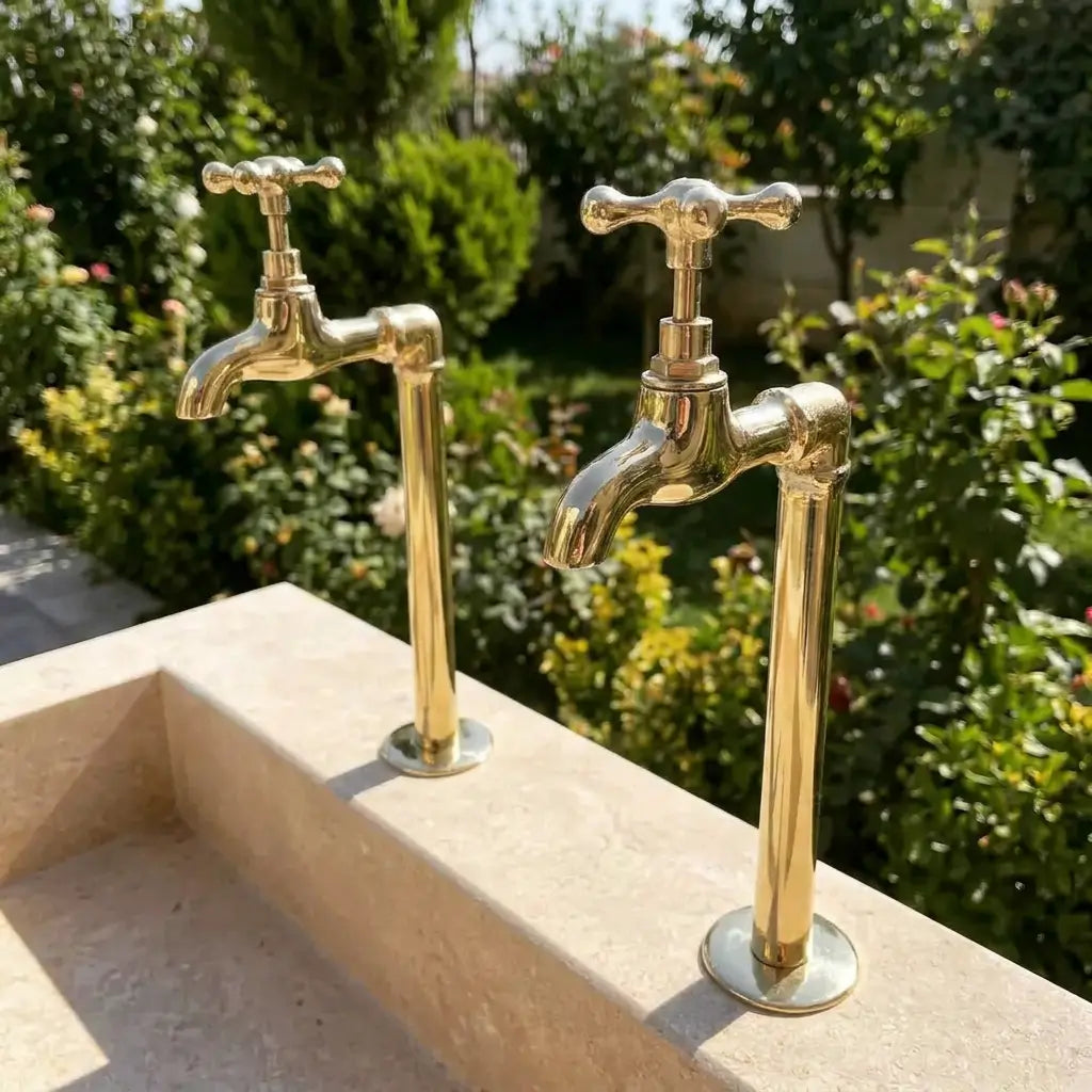A pair of unlacquered brass wall mount water faucets with a vintage Moroccan inspired design, installed outdoors on a stone sink against a garden background.