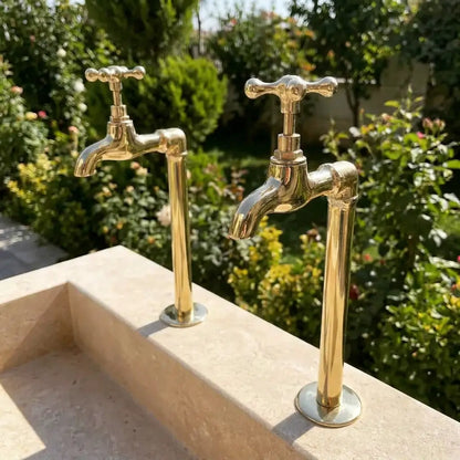 A pair of unlacquered brass wall mount water faucets with a vintage Moroccan inspired design, installed outdoors on a stone sink against a garden background.