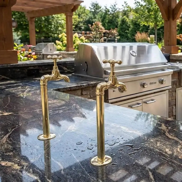 Two elegant brass water filter faucets installed on a luxury dark marble countertop next to a stainless steel grill in an outdoor kitchen setup.