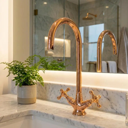 Polished copper gooseneck faucet with dual cross handles on a white marble vanity top in front of a large bathroom mirror.