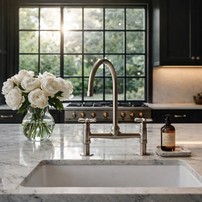 Elegant warm brushed nickel Heritage bridge kitchen faucet mounted on a polished white marble countertop with a vase of white roses and classic grid window background.