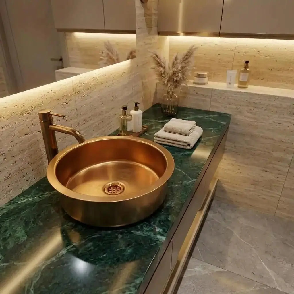 Top view of a round brass vessel sink with a brushed bronze faucet on a green marble countertop with a travertine stone wall and LED strip lighting