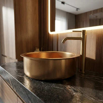 Side view of a handcrafted round brass vessel basin with a matching tall modern faucet, installed on a dark veined granite countertop with a backlit mirror.
