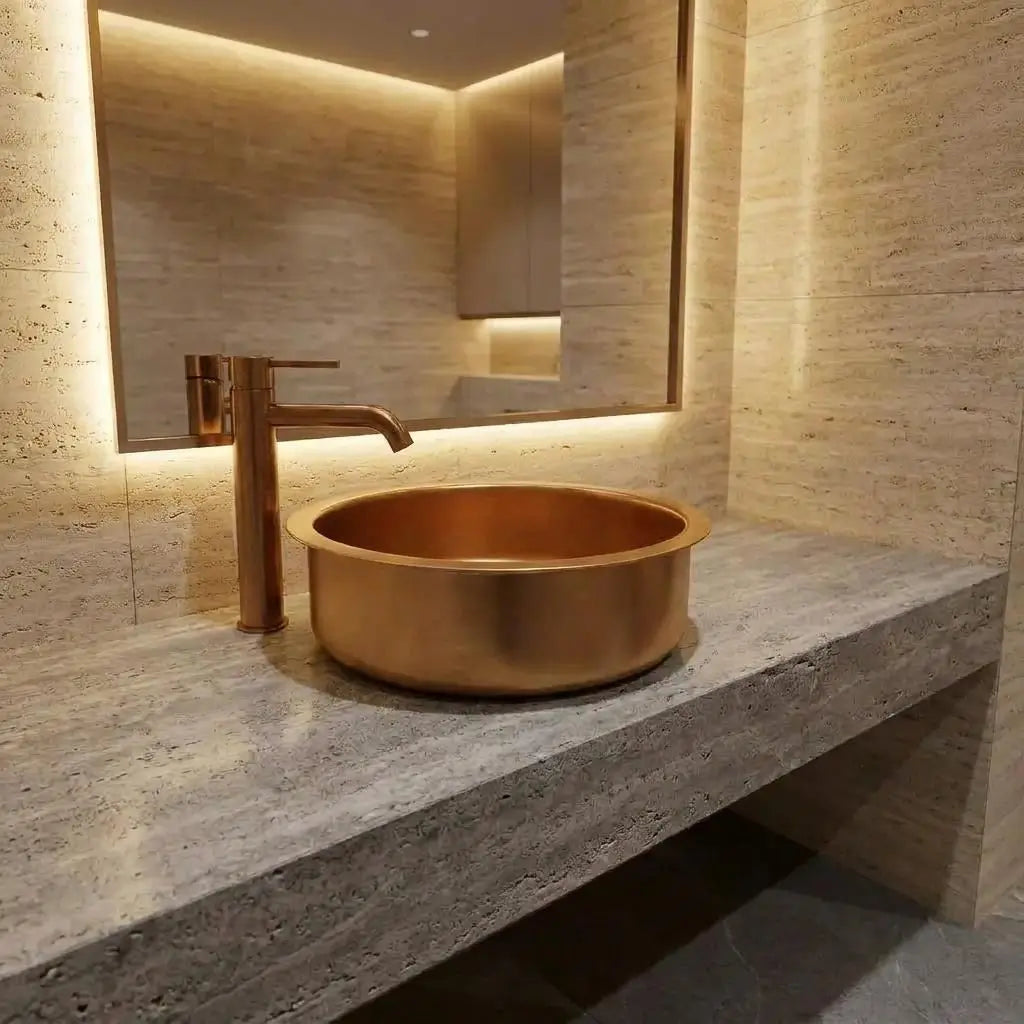 Handcrafted round brass vessel sink with a brushed finish, installed on a grey stone countertop with a matching modern brass faucet in a luxury bathroom setting.