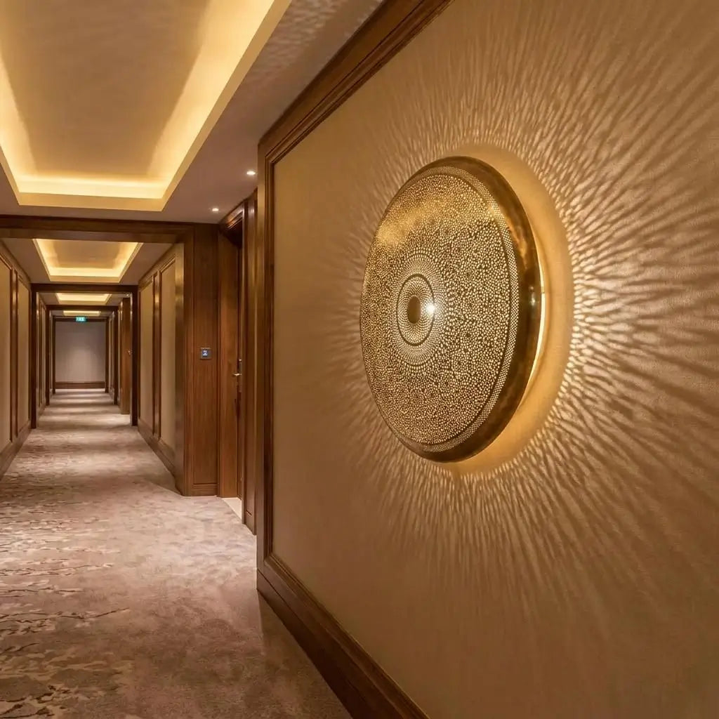 Round perforated brass wall sconce creating a starburst light effect on a long luxury hotel corridor wall.