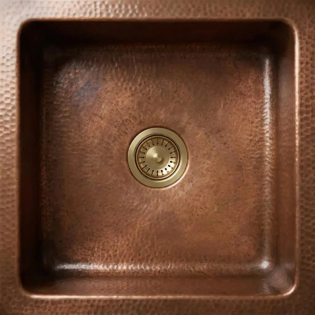 Top-down view of a solid brass kitchen sink waste drainer installed inside a square hammered copper basin.