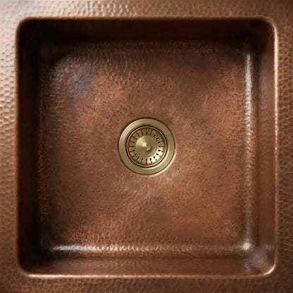 Top-down view of a solid brass kitchen sink waste drainer installed inside a square hammered copper basin.