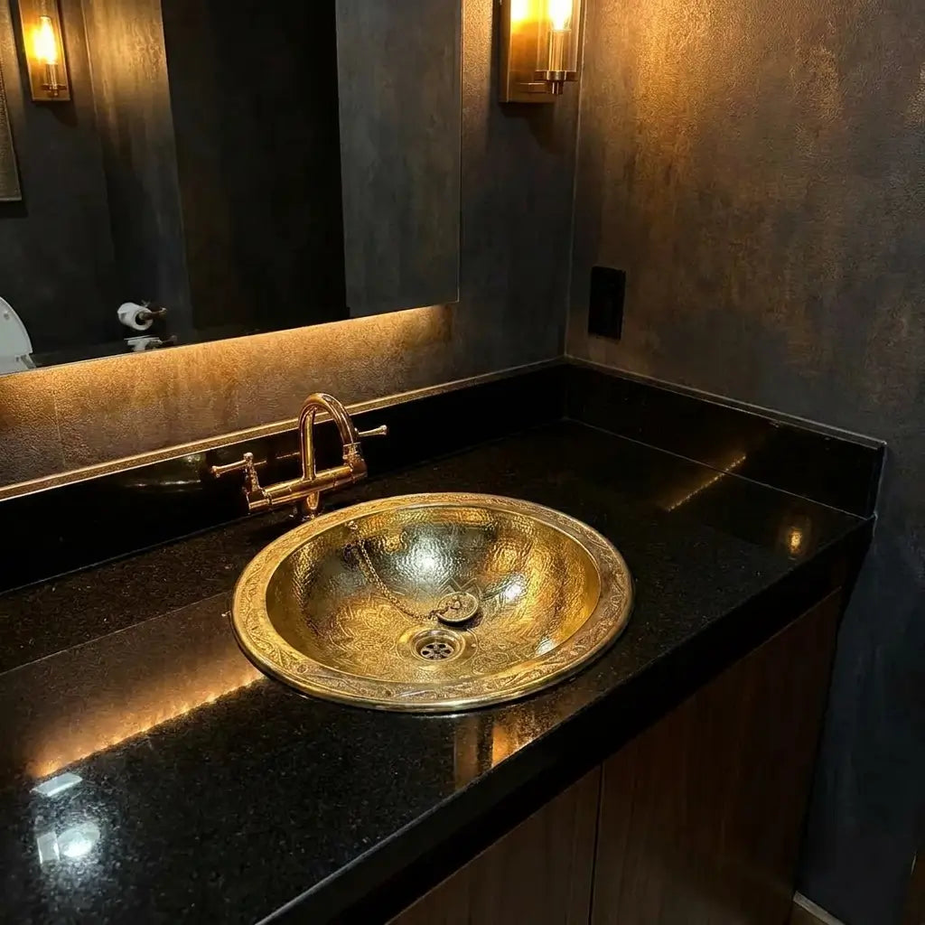 Polished round brass vessel sink with a hammered finish set on a dark granite countertop under a backlit mirror in a moody restroom.