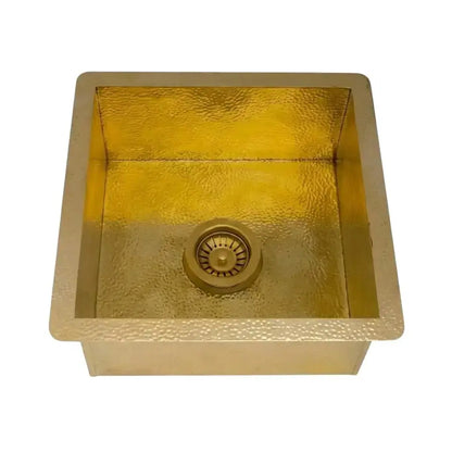 Isolated square hammered brass kitchen sink with integrated drain assembly displayed on a clean solid white studio background.