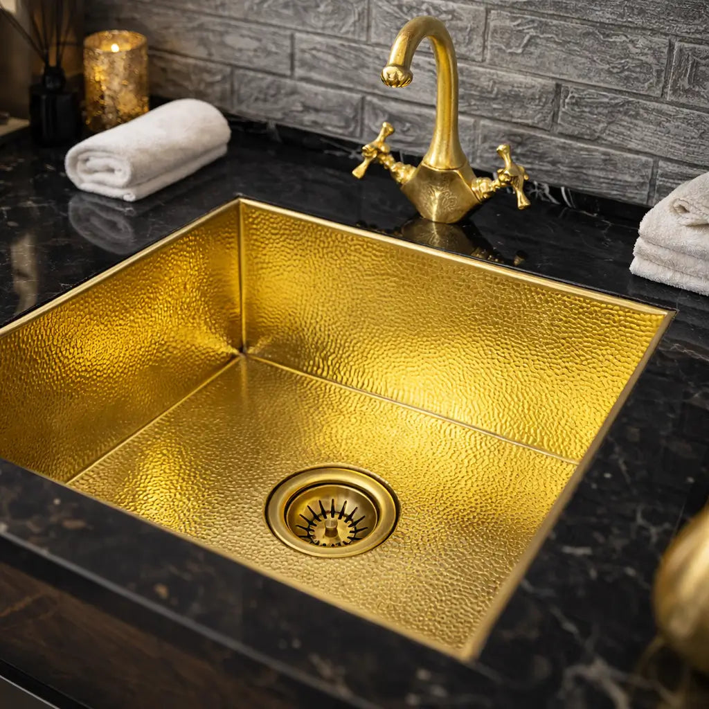 A square undermount brass Moroccan sink with a hammered interior, installed in a dark marble countertop with a vintage gold faucet, a rolled white towel, and a textured grey brick wall.