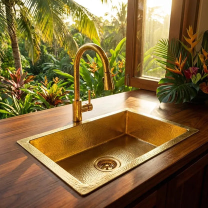Golden brass undermount sink installed on a rich wooden countertop next to an open window overlooking a lush tropical garden