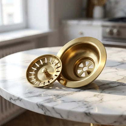 An unlacquered brass 3.5 inch kitchen sink waste flange and its matching removable strainer basket resting on a white marble tabletop.