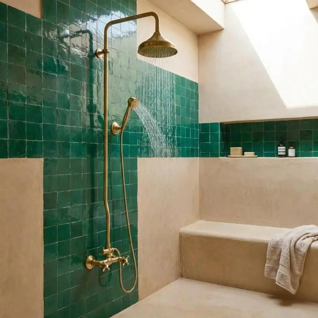 Unlacquered brass exposed pipe shower system with flowing water from the rainfall head and handheld sprayer, mounted on a green zellige tile wall inside a modern plastered bathroom alcove.